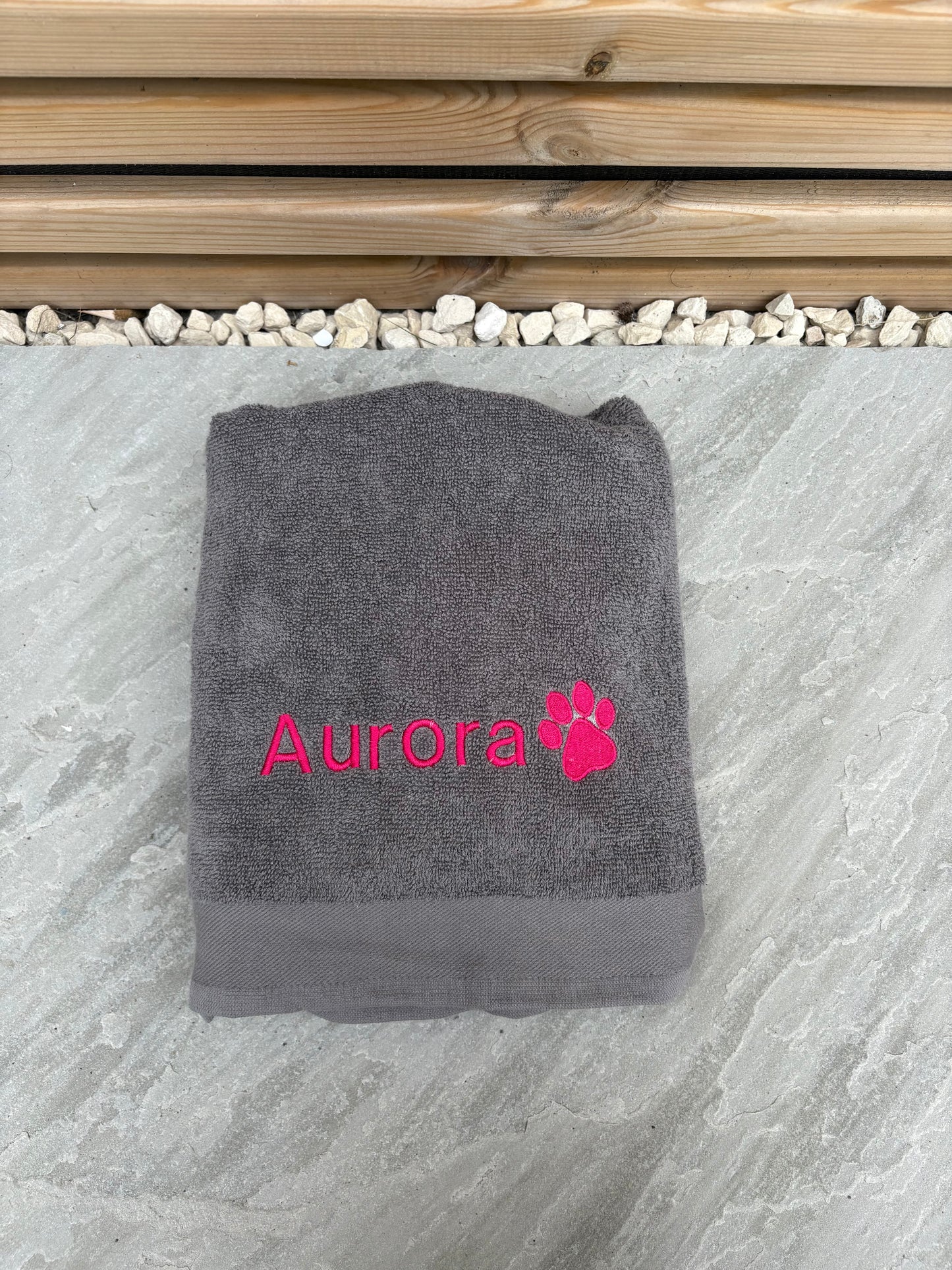 Gray towel with 'Aurora' and a paw print in pink on a stone surface.