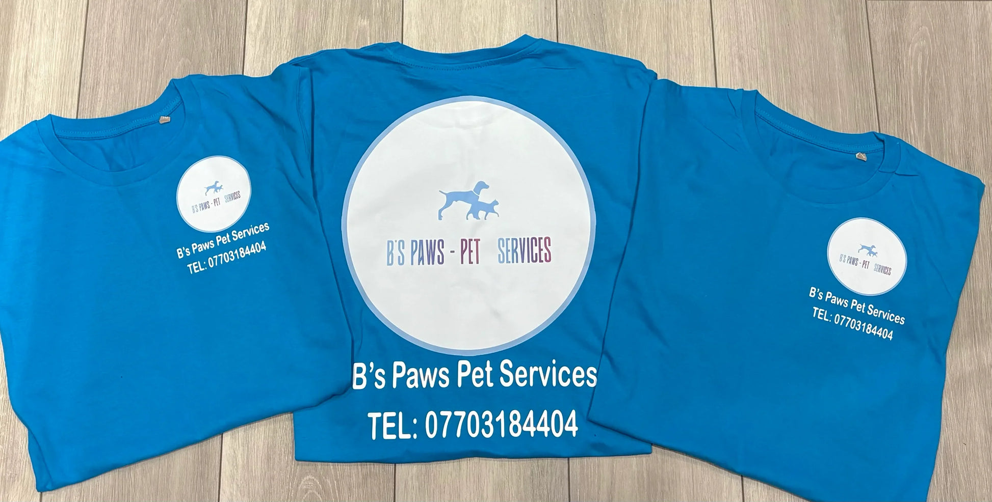 Three blue t-shirts with 'B's Paws Pet Services' branding on a wooden surface