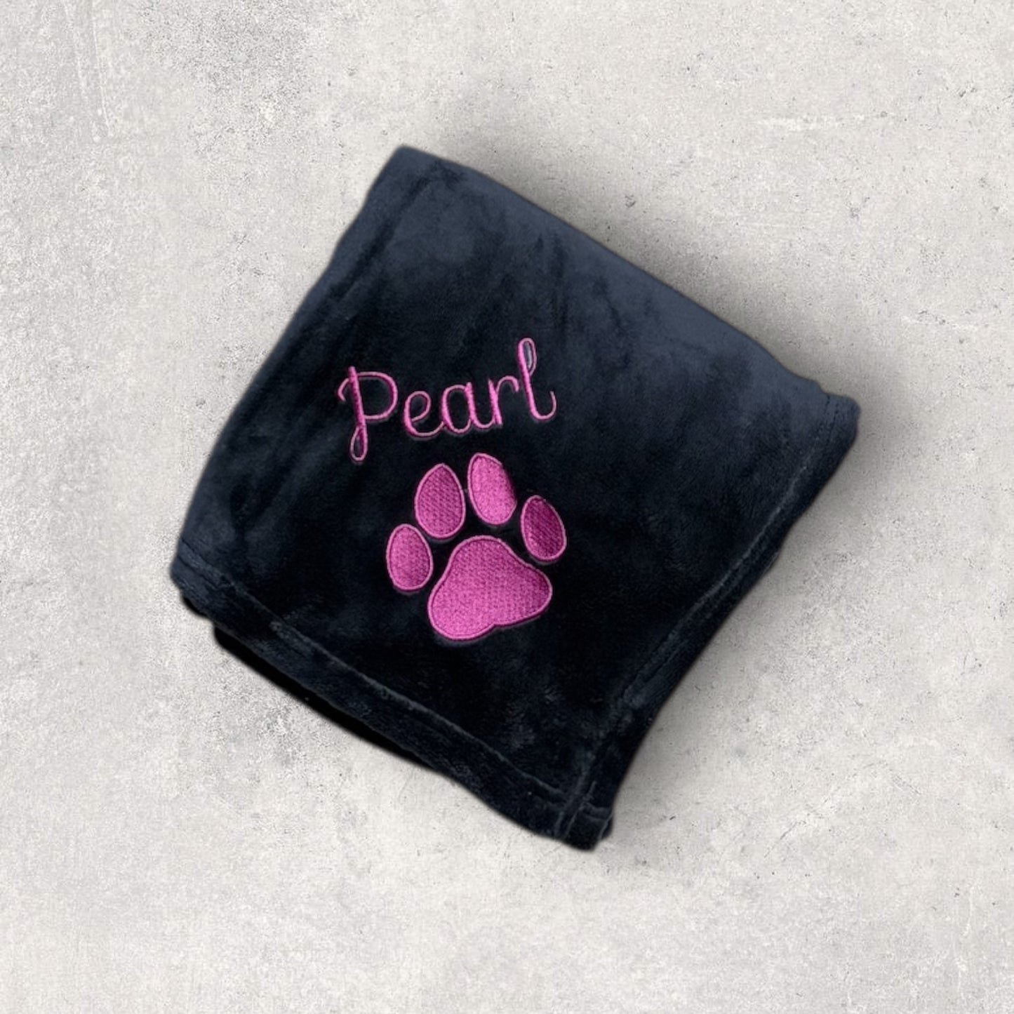 Black blanket with pink paw print and 'Pearl' text on a gray surface