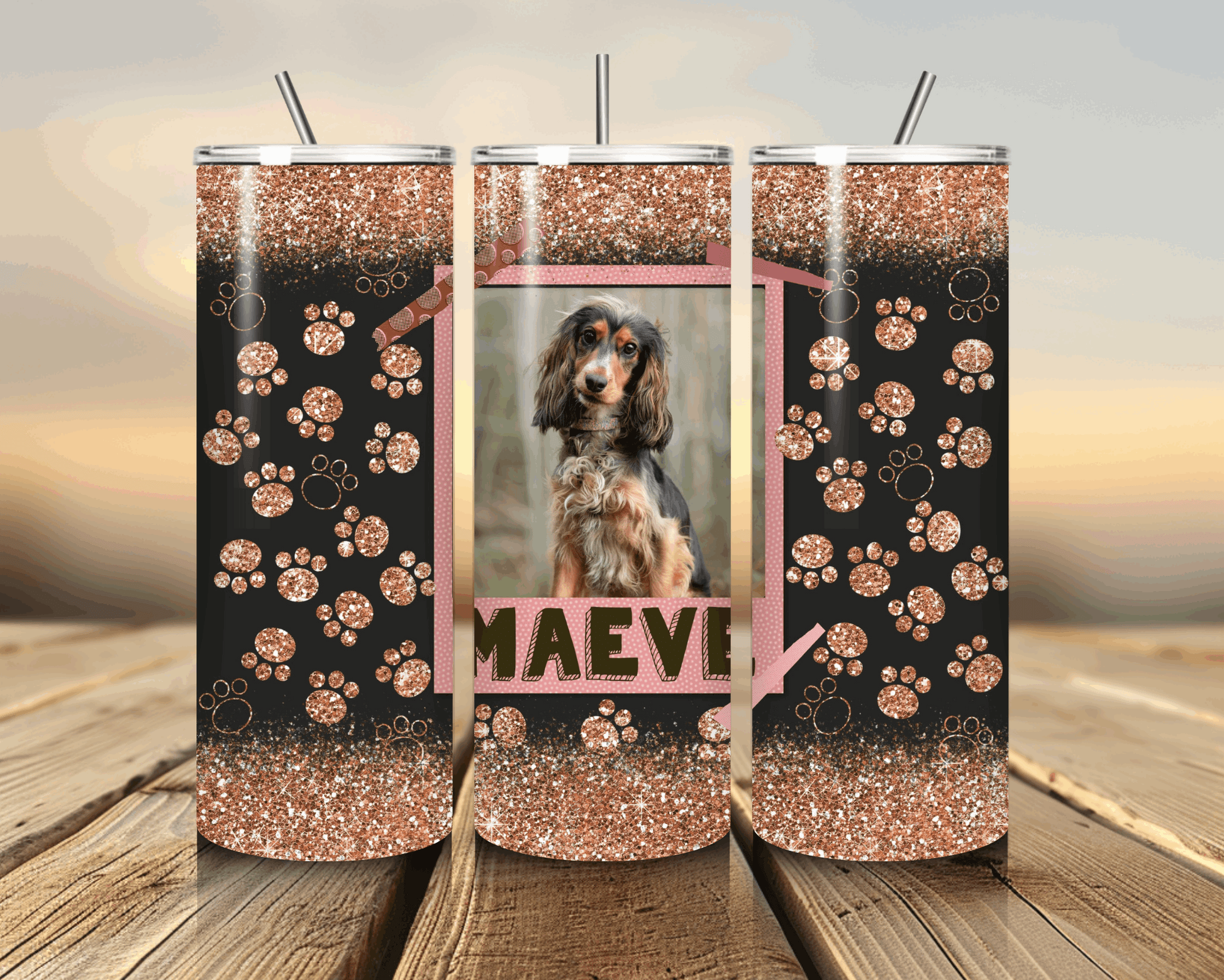 Three glittery tumblers with paw patterns and a photo of a dog named Maeve on a wooden surface.