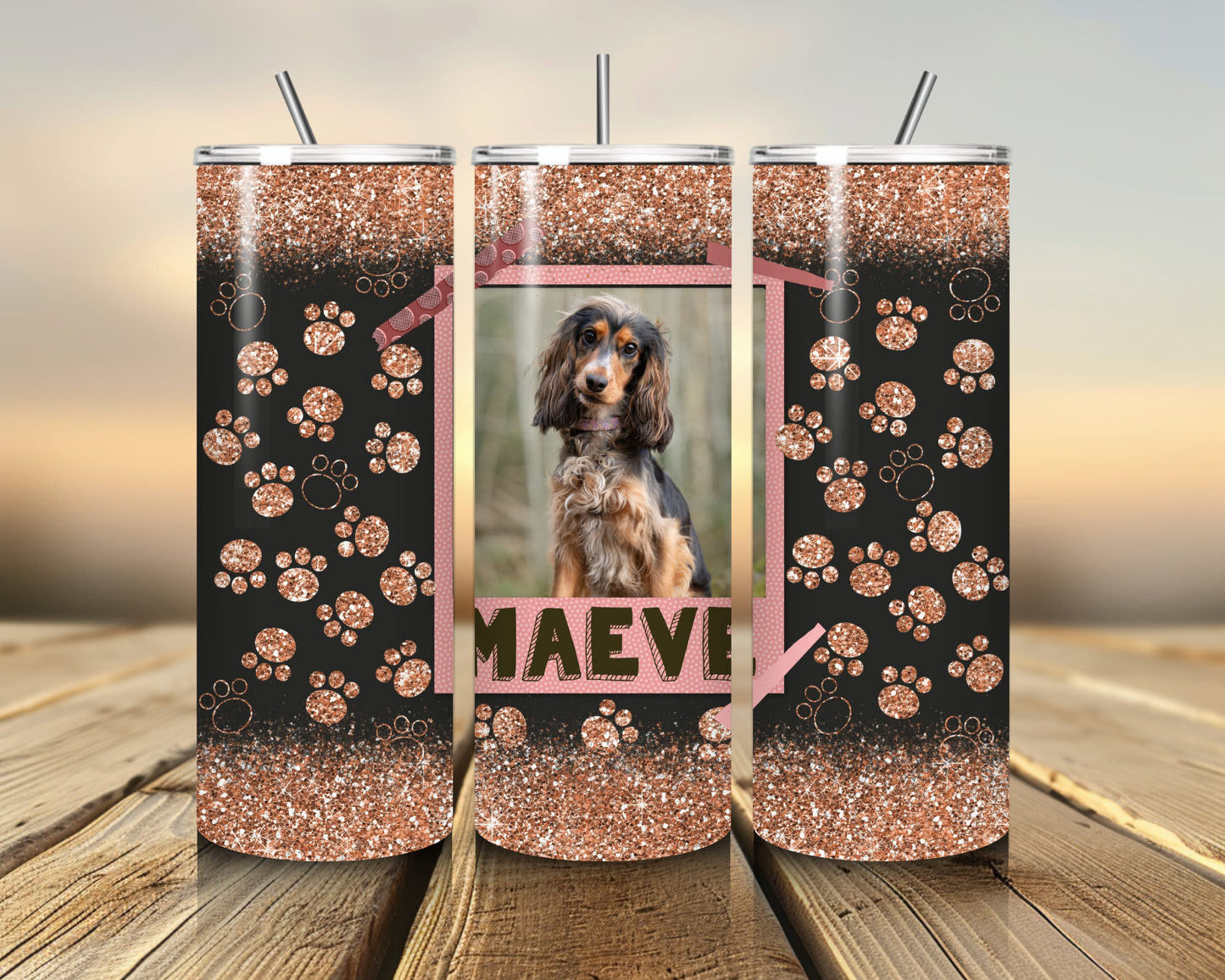 Three glittery tumblers with paw patterns and a photo of a dog named Maeve on a wooden surface.