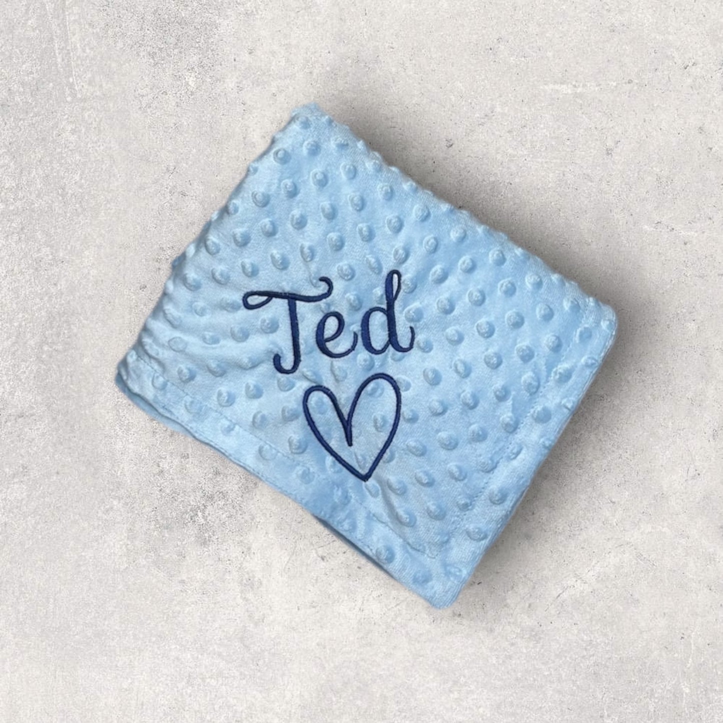 Light blue textured blanket with 'Ted' and a heart embroidered on it, placed on a light gray surface.