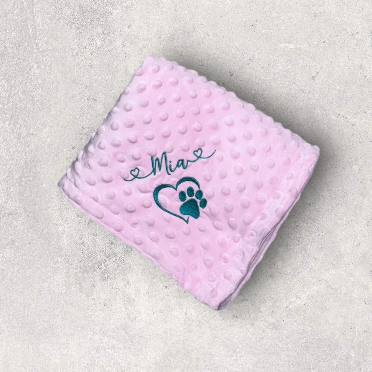 Pink blanket with 'Mia' and a paw print on a gray surface