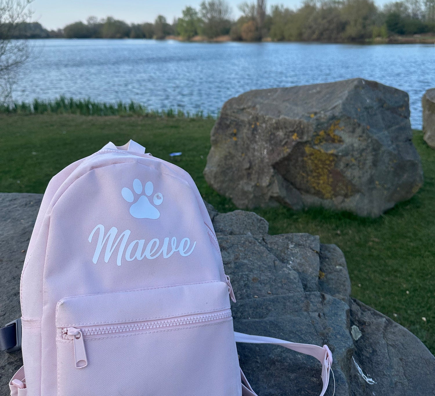 Pink backpack with 'Maeve' and a paw print on it, placed outdoors near a lake and rocks.